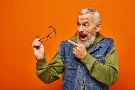 joyous mature man in vibrant attire with beard holding his glasses in hand on orange backdrop