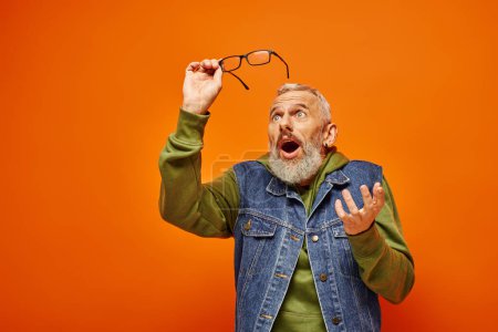 good looking surprised mature man in vivid attire holding glasses and posing on orange backdrop