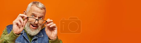 Foto de Alegre hombre maduro barbudo gris en sudadera con capucha vibrante y chaleco de mezclilla posando sobre fondo naranja, bandera - Imagen libre de derechos
