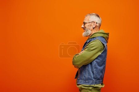 handsome merry mature man in green hoodie and denim vest posing in profile on orange backdrop