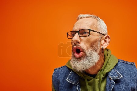 gut aussehender reifer Mann in grünem Kapuzenpulli mit Brille posiert mit offenem Mund auf orangefarbenem Hintergrund