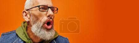 handsome mature man in green hoodie with beard posing with open mouth on orange backdrop, banner
