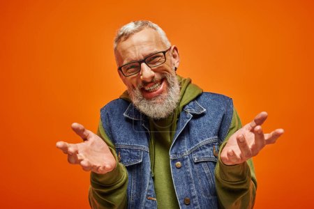 Foto de Alegre hombre guapo con barba y gafas encogiéndose de hombros y sonriendo a la cámara sobre fondo naranja - Imagen libre de derechos