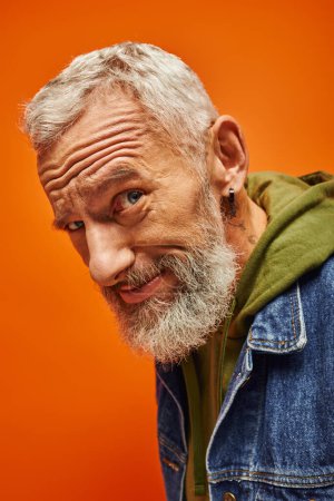 beau modèle masculin joyeux avec barbe grise posant sur fond orange et souriant à la caméra
