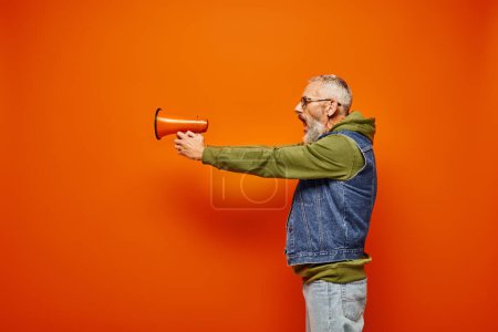 Photo for Handsome mature male model in green hoodie and stylish denim vest aiming with megaphone in hands - Royalty Free Image