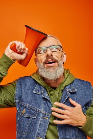 Photo for Joyous surprised mature man in green hoodie and denim vest posing with megaphone on orange backdrop - Royalty Free Image