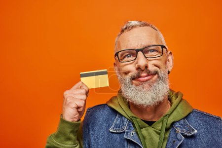 Photo for Cheerful good looking mature man in vibrant clothes with glasses holding credit card near his face - Royalty Free Image