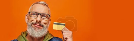 jolly attractive mature man in vivid clothes with glasses holding credit card near his face, banner