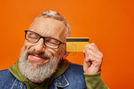 Photo for Handsome mature male model in vibrant clothing posing with closed eyes and holding credit card - Royalty Free Image
