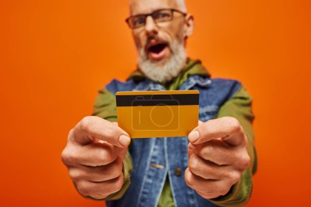 Foto de Centrarse en la tarjeta de crédito en manos de hombre barbudo desenfocado maduro en traje vibrante sobre fondo naranja - Imagen libre de derechos