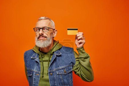 Photo for Jolly attractive mature man in denim vest with glasses looking at credit card on orange backdrop - Royalty Free Image