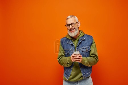 Téléchargez les photos : Joyeux beau modèle masculin mature en tenue vibrante tenant smartphone et souriant à la caméra - en image libre de droit