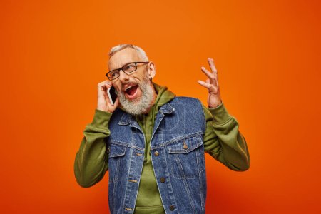 Photo for Good looking joyful mature man in vibrant green hoodie and denim vest with glasses talking by phone - Royalty Free Image