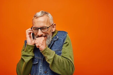 cheerful handsome mature man in trendy denim vest with beard and glasses talking happily by phone