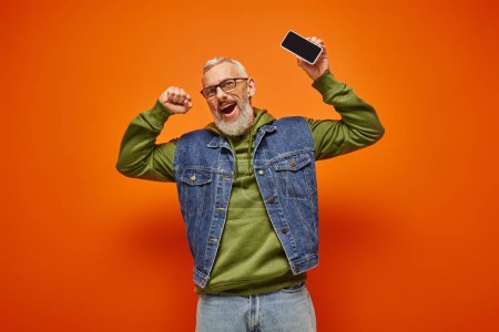 Photo for Joyful attractive mature man in green hoodie and denim vest holding phone and looking at camera - Royalty Free Image