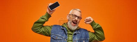 Photo for Jolly handsome mature man in green hoodie and denim vest holding phone and looking at camera, banner - Royalty Free Image
