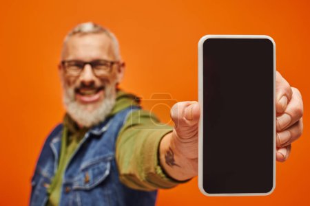 Foto de Se centran en el teléfono inteligente en manos del hombre maduro alegre borrosa en ropa vibrante sobre fondo naranja - Imagen libre de derechos