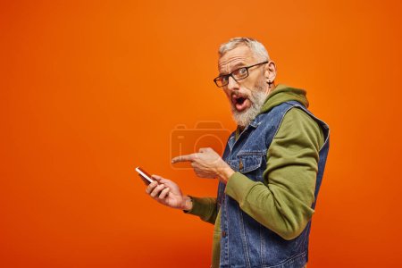 Photo for Surprised handsome mature man in green hoodie pointing at smartphone and looking at camera - Royalty Free Image