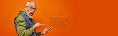 Photo for Shocked attractive mature man in green hoodie pointing at smartphone and looking at camera, banner - Royalty Free Image