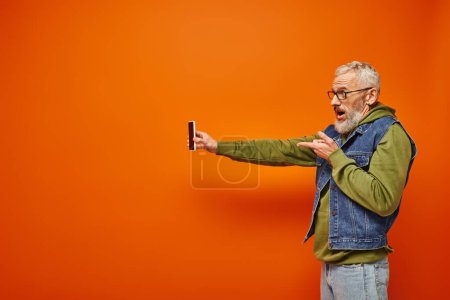 Photo for Good looking mature male model in vibrant clothes with beard and glasses holding smartphone - Royalty Free Image
