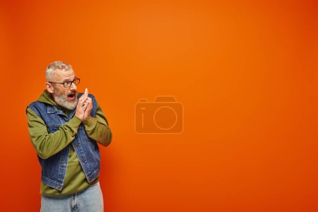 Photo for Attractive mature male model in green hoodie and denim vest talking by smartphone on orange backdrop - Royalty Free Image