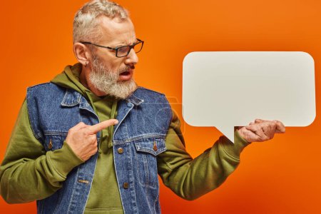 Photo for Confused handsome mature man in denim vest looking at speech bubble in his hand on orange backdrop - Royalty Free Image