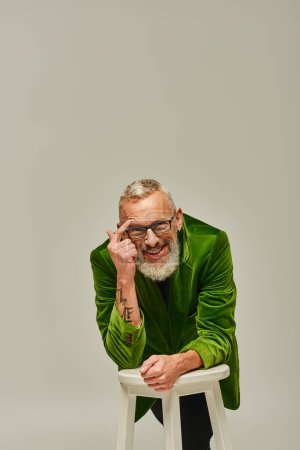Photo for Joyous bearded mature man in vivid attire with tattoos posing with tall chair and smiling at camera - Royalty Free Image