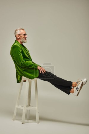 Photo for Handsome funky mature male model in bright attire with accessories posing on tall chair in profile - Royalty Free Image