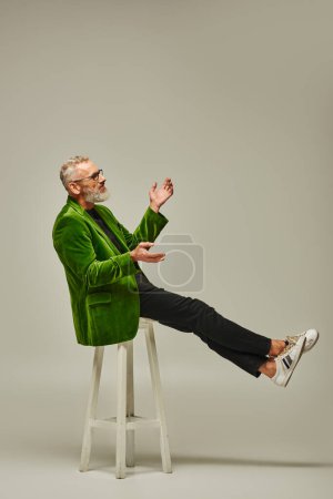 joyous mature man in green blazer with glasses sitting on tall chair in profile on beige backdrop