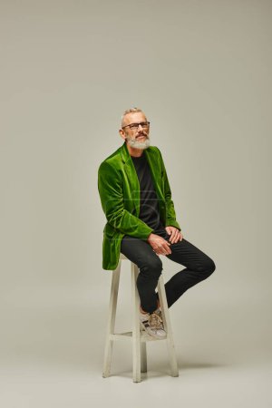 Photo for Good looking mature male model with beard and glasses sitting on tall chair and looking at camera - Royalty Free Image