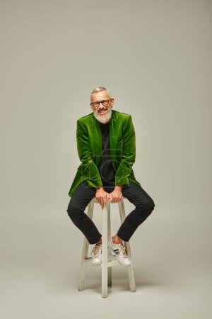 cheerful funky male model with beard and glasses sitting on tall chair and smiling at camera