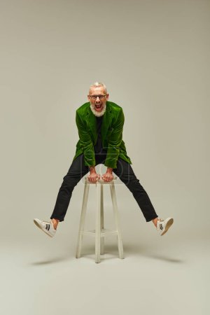 joyful good looking mature man in vibrant attire posing on tall chair actively and smiling at camera
