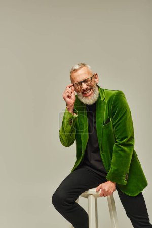 Foto de Bien parecido hombre funky en chaqueta verde sentado en silla alta y sonriendo sinceramente con los ojos cerrados - Imagen libre de derechos