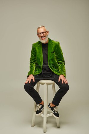 Photo for Cheerful bearded attractive mature man in green blazer sitting on tall chair and smiling at camera - Royalty Free Image