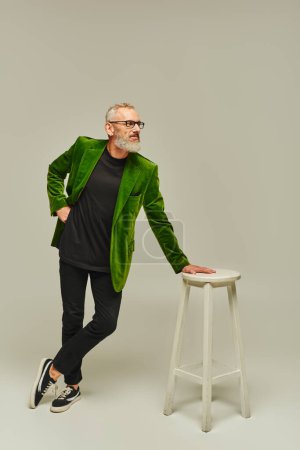 good looking mature man with beard in vibrant attire posing next to tall chair and looking away