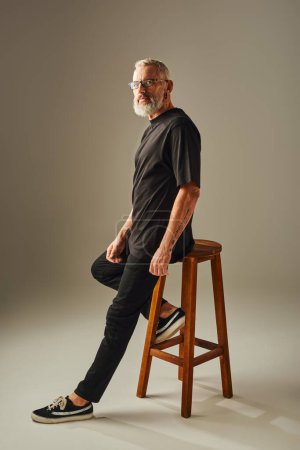 handsome mature man in black t shirt sitting on tall chair and looking at camera on beige backdrop