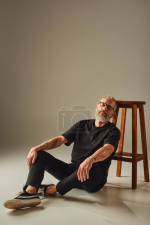attraktiver, reifer Mann in schwarzem T-Shirt mit Bart sitzt auf dem Boden in der Nähe eines hohen Stuhls mit geschlossenen Augen
