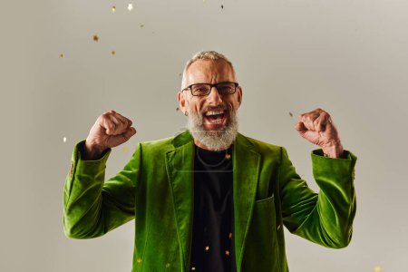 Photo for Handsome jolly mature man in vibrant blazer showing fists under confetti rain on beige backdrop - Royalty Free Image