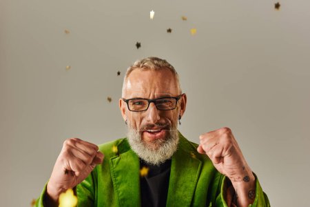Photo for Joyous good looking mature male model in vibrant blazer showing fists at camera under confetti rain - Royalty Free Image