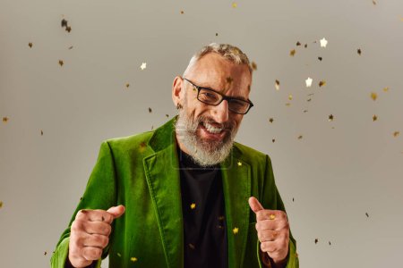 Photo for Joyful good looking mature man in green blazer looking at camera under confetti on beige backdrop - Royalty Free Image