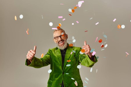Photo for Joyful mature male model in green blazer showing thumbs up under confetti rain on beige background - Royalty Free Image
