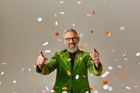 Photo for Joyous handsome mature man in green blazer pointing at camera under confetti rain on beige backdrop - Royalty Free Image