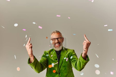 Téléchargez les photos : Joyeux homme mûr en vert vibrant blazer avec des lunettes montrant les doigts du milieu et souriant à la caméra - en image libre de droit