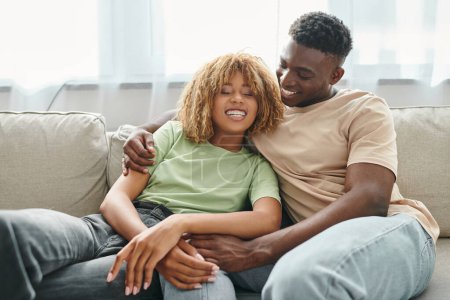 Photo for Cheerful black man embracing happy girlfriend in braces while sitting on cozy couch in living room - Royalty Free Image