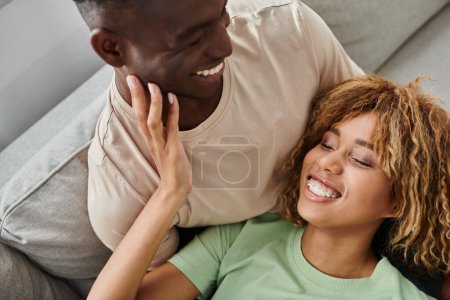 african american couple laughing while resting on sofa, cheerful woman lying on laps of boyfriend