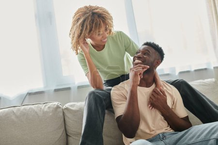 freudige afrikanisch-amerikanische Frau schaut ihren Freund im Wohnzimmer an, schwarzes Paar und hochwertige Zeit