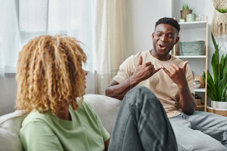 Foto de Alegre hombre negro sonriendo mientras se comunica con el lenguaje de señas con la novia en la sala de estar - Imagen libre de derechos