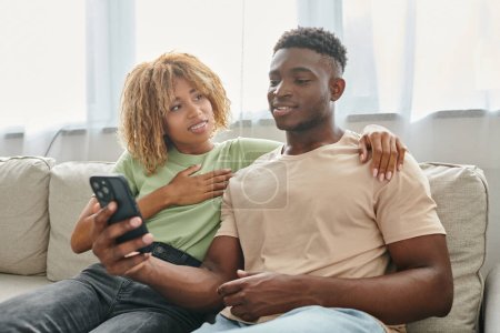 szczęśliwy Afryki amerykanka kobieta w aparat na zęby przytulanie i patrząc na mężczyznę z smartphone, komunikacja