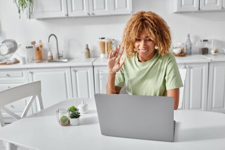 femme afro-américaine souriante utilisant le langage des signes lors d'un appel vidéo sur ordinateur portable, agitant la main