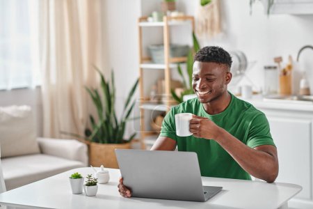 Hombre negro sonriente disfrutando de una taza de café mientras usa el portátil en la cocina, freelancer en casa
